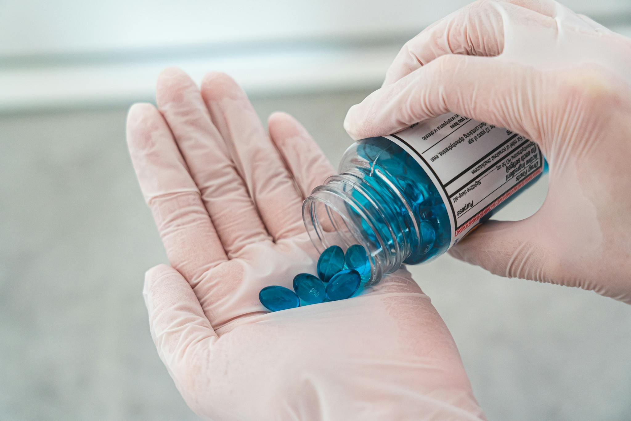 Gloved hands carefully pouring blue oval pills from a prescription bottle.